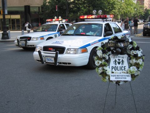 1991 Ford LTD Crown Victoria - Two white modern NYPD cruisers parked on a city street beside a floral memorial wreath reading Police Radio Motor Patrol Association.