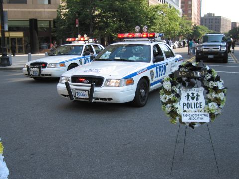 1991 Ford LTD Crown Victoria - White NYPD patrol cars parked downtown beside a Lest We Forget police memorial wreath on an easel.