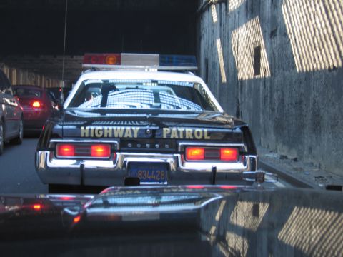 1991 Ford LTD Crown Victoria - Rear view of a black Dodge Highway Patrol cruiser with caged windows and brake lights glowing in dim tunnel traffic.