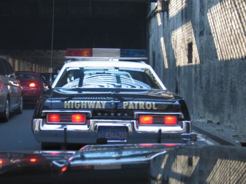 1991 Ford LTD Crown Victoria - Rear of a black Dodge Highway Patrol sedan with lightbar and plate 834428, brake lights lit in tunnel traffic beside a stone wall.