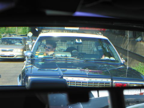 1991 Ford LTD Crown Victoria - Framed by a car's rear window, a sunglasses-wearing driver sits in a vintage Chrysler police cruiser amid slow-moving traffic.
