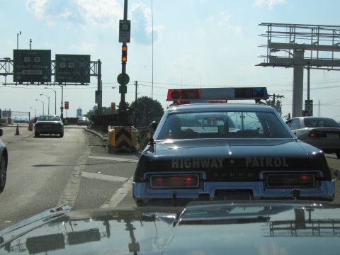 1991 Ford LTD Crown Victoria - Rear of a black Dodge Highway Patrol sedan with lightbar stopped at a traffic signal near overhead highway direction signs.