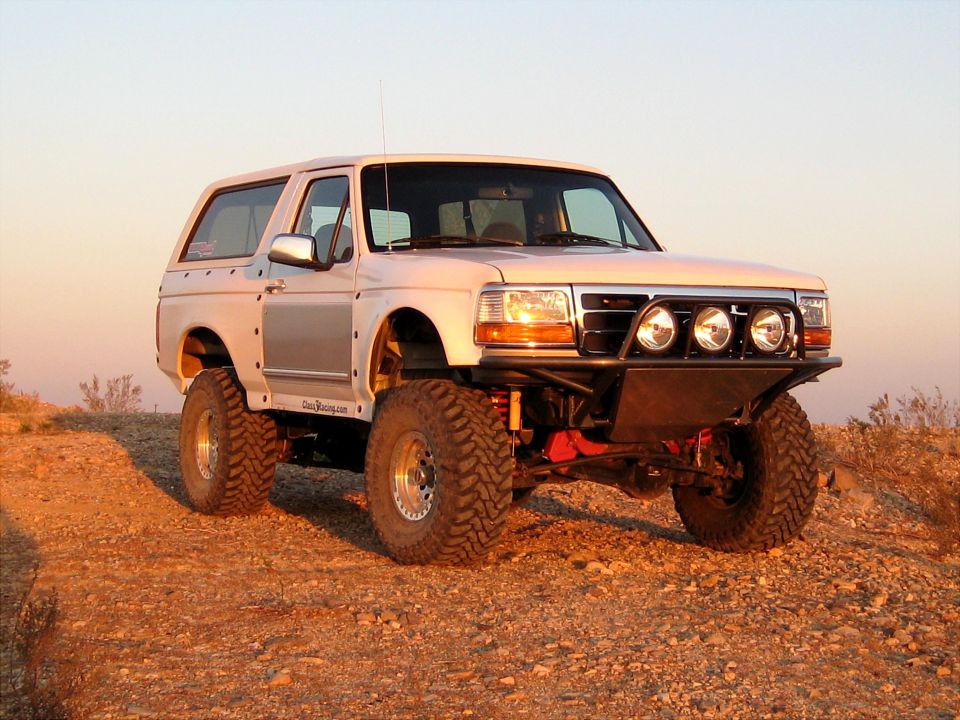 White 1996 Ford Bronco with off-road modifications, including a custom bumper and large tires, parked on a dirt hill