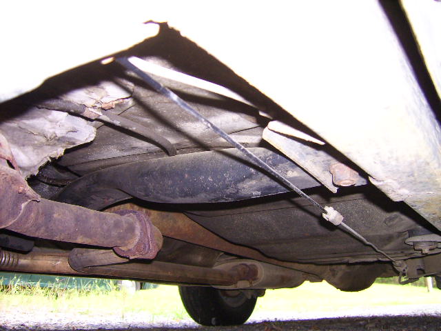 Underside of a car showing rusty exhaust pipe, muffler, torn heat shield and routed brake cable