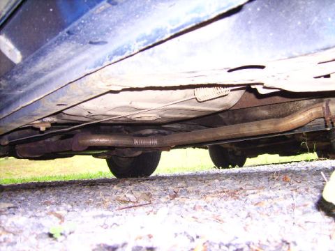 1982 Ford LTD Crown Victoria - Ground-level view under the passenger side of a dark blue car, looking up at the exhaust system: a surface-rusted pipe with a flex/braided…