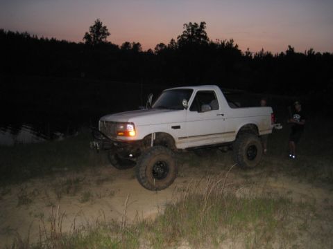 1996 Ford Bronco - A lifted white Ford Bronco is parked on a dirt path next to a body of water at dusk. Two people are standing behind the vehicle.