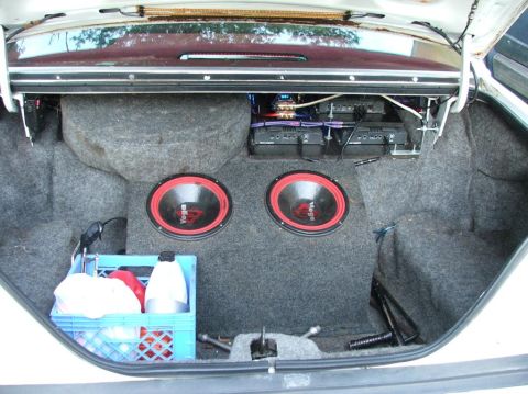 1985 Ford LTD Crown Victoria - The trunk of a white sedan is open, revealing a custom car audio system with two subwoofers and two amplifiers. A blue plastic crate contai…