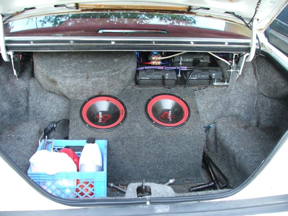 Trunk of a white sedan with custom car audio system, two subwoofers, and amplifiers.