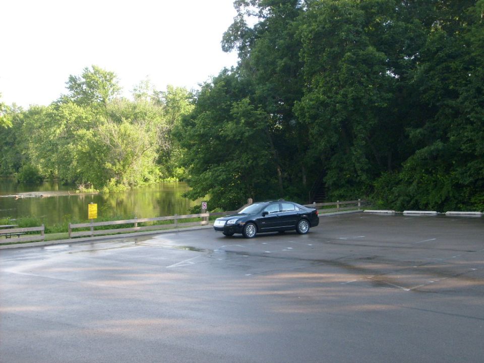 Black sedan parked in a lot near water and trees.