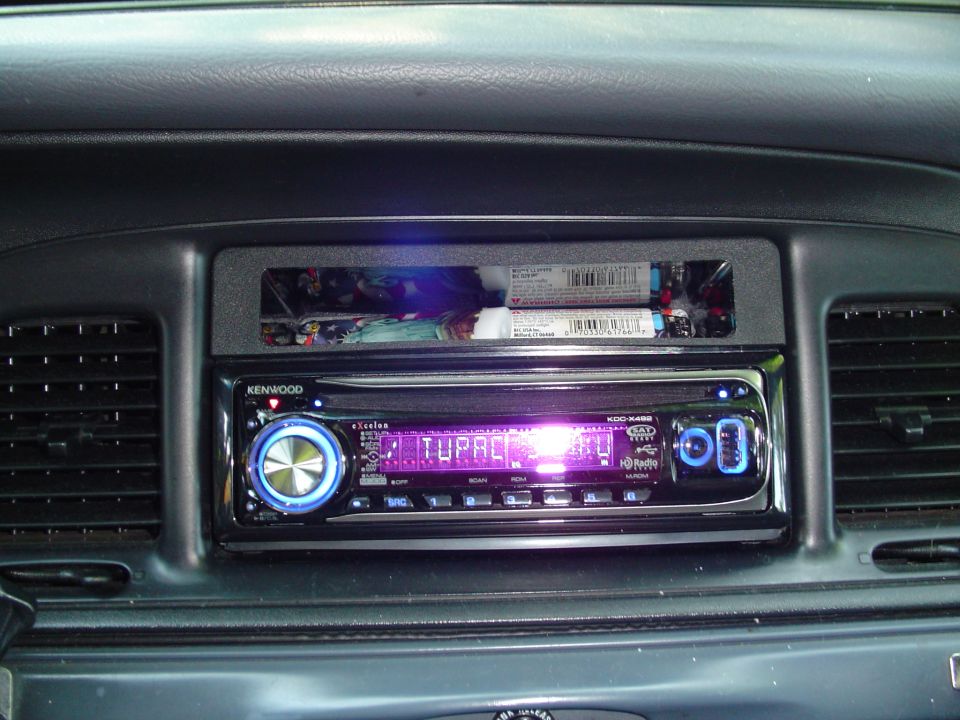 Car dashboard with a Kenwood stereo displaying 'TUPAC' and a storage compartment above it