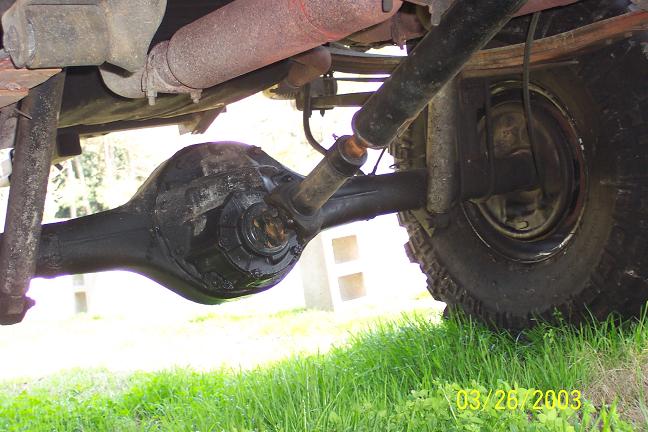 Underside view of a vehicle's rear axle, differential, driveshaft, and tire, with grass in the foreground