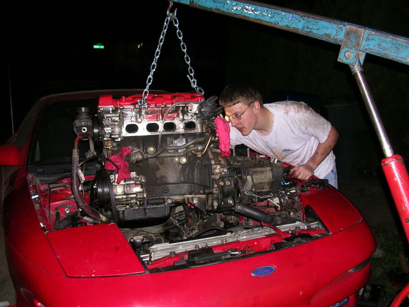Man in glasses looking at a red car engine being lifted by a chain hoist. The car's hood is open