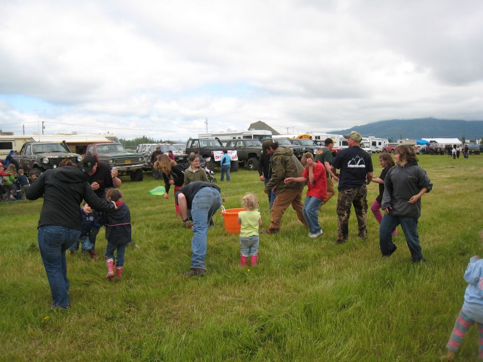 People gathered in a grassy field with several SUVs and RVs parked in the background under a cloudy sky