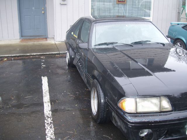 Black Ford Mustang with aftermarket wheels parked on a wet asphalt lot