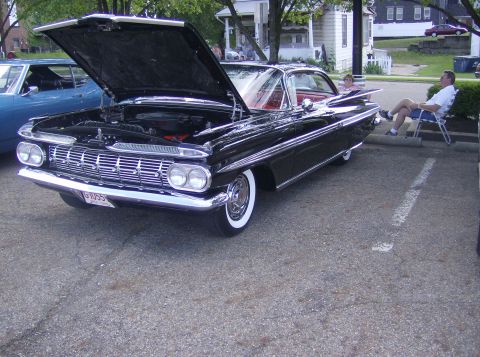 1970 Ford Ranchero - A classic black 1959 Chevrolet Impala is displayed with its hood open, while a man relaxes in a chair beside it.