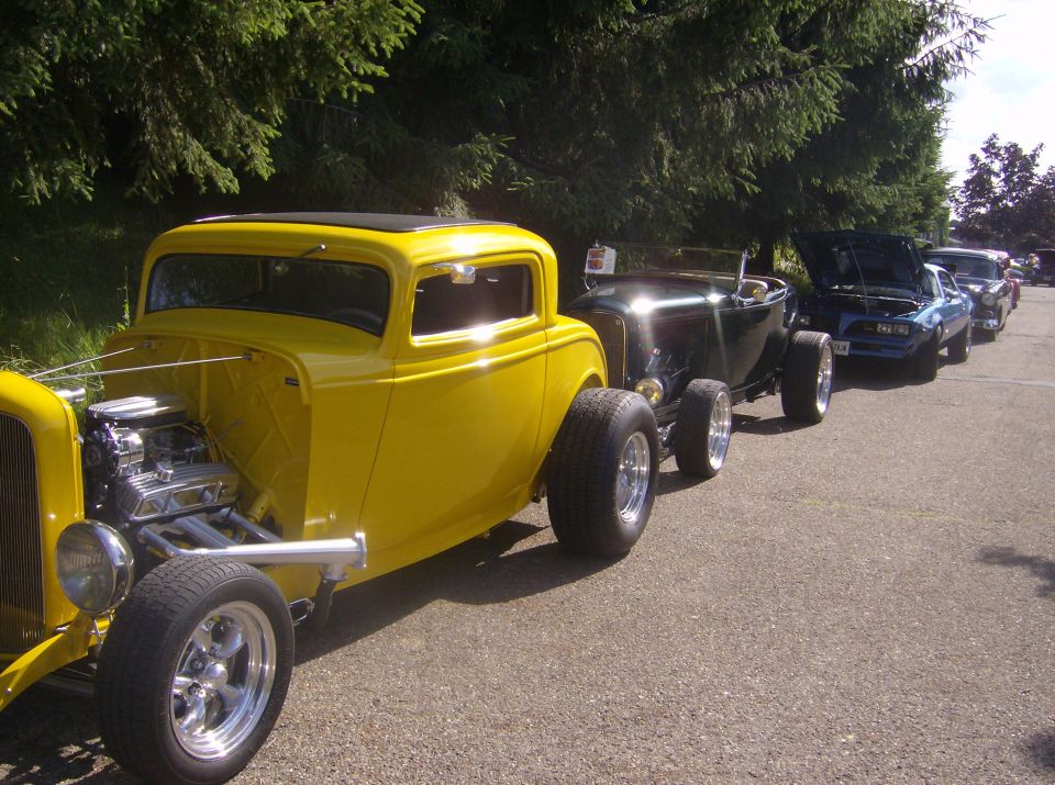 Yellow hot rod in front, followed by black convertible and blue car. Parked on asphalt with trees