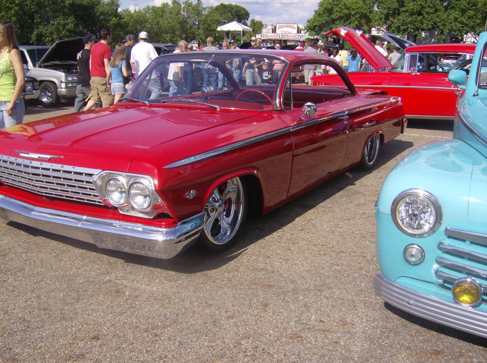 Red 1962 Chevrolet Impala lowrider with chrome wheels, parked next to a blue car