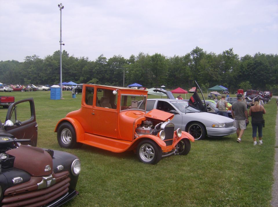 Orange vintage hot rod with open hood, parked on grass at a car show.