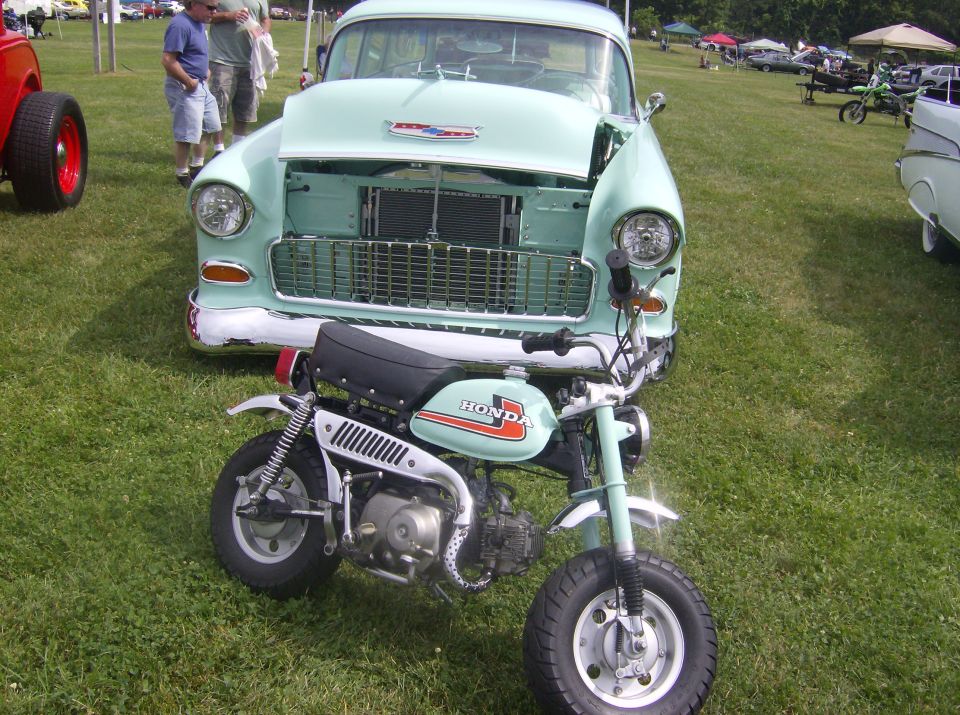 Mint green Honda Z50 mini-bike in front of a light blue 1955 Chevrolet Bel Air with hood open.