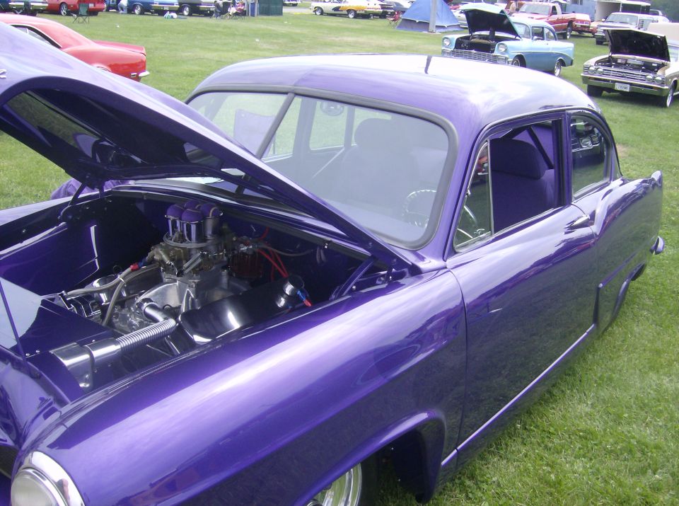 Custom purple vintage sedan with open hood showing chrome engine, parked on grass.