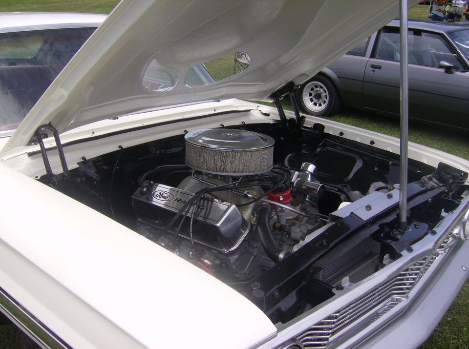 White classic Ford with open hood showing engine. Chrome air cleaner and Ford Racing valve covers visible. Gray sedan in background.