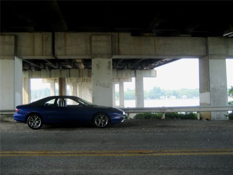 1993 Ford Probe - A dark blue coupe is parked under a concrete overpass, facing right, with a body of water and trees visible in the background.