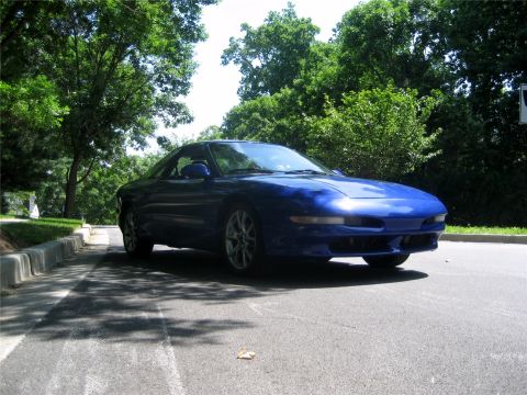 1993 Ford Probe - A blue Ford Probe is parked on the side of a paved road, with lush green trees and foliage in the background. The car is angled towards the…