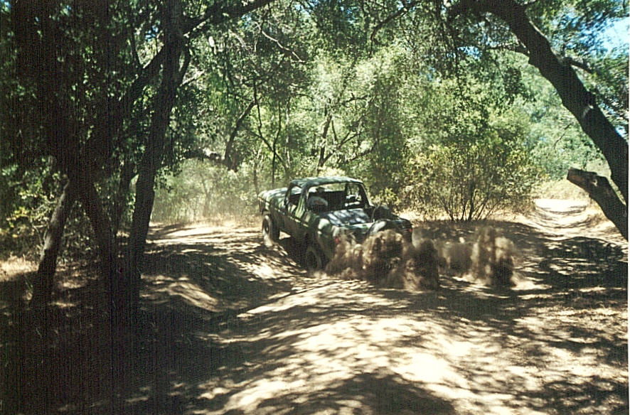 Dark green topless Bronco kicking up dust on a shaded dirt trail through oak trees