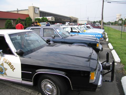 1991 Ford LTD Crown Victoria - row of classic police cars parked in a lot, led by a black-and-white Highway Patrol sedan with push bar, others in white and blue behind.