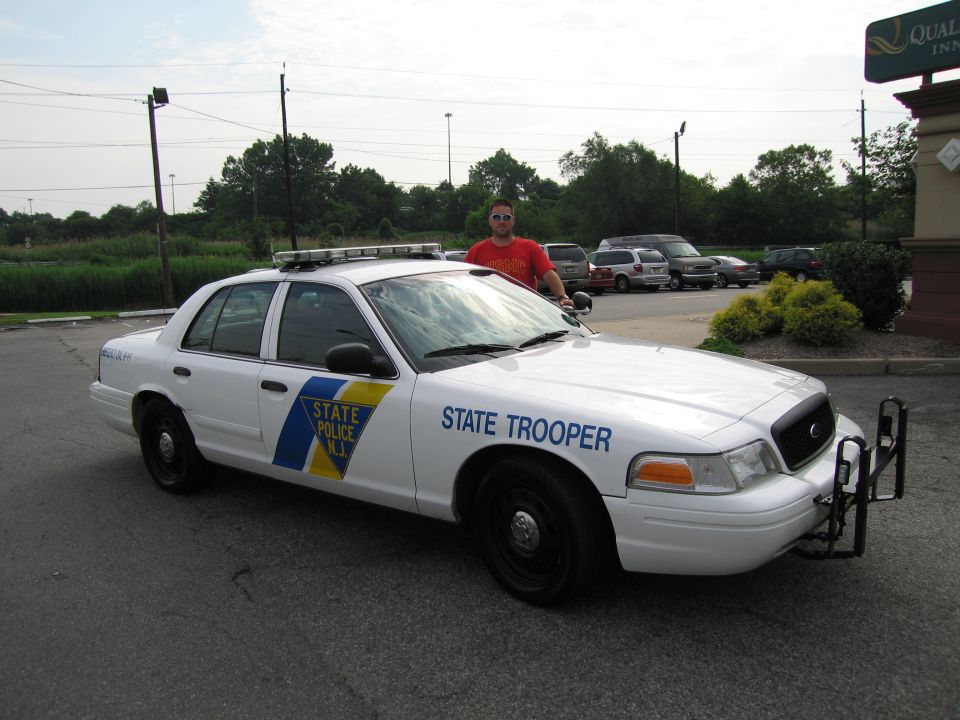 Man in red shirt standing beside white NJ State Police Crown Victoria patrol car