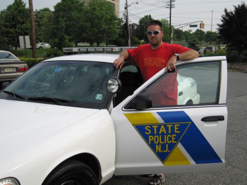 1991 Ford LTD Crown Victoria - man wearing a red USMC shirt and sunglasses leans on the open driver door of a white New Jersey State Police patrol car.