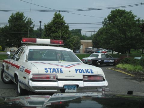 1991 Ford LTD Crown Victoria - vintage 1970s white State Police sedan with red-and-white roof light bar and vanity plate PURSUT, with a dark older patrol car behind.