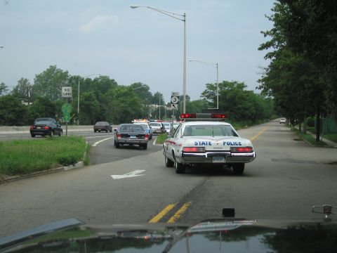 1991 Ford LTD Crown Victoria - white vintage State Police sedan with roof light bar drives on a road, escorting a line of other police cars under a street lamp.
