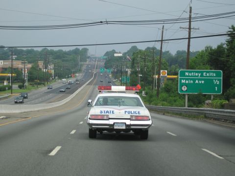1991 Ford LTD Crown Victoria - white vintage State Police sedan with roof light bar travels a multi-lane highway near a green Nutley Exits / Main Ave sign.