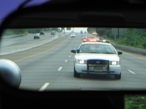1991 Ford LTD Crown Victoria - Seen through a rear window, a white Ford Crown Victoria police car with lights follows on a multi-lane highway.