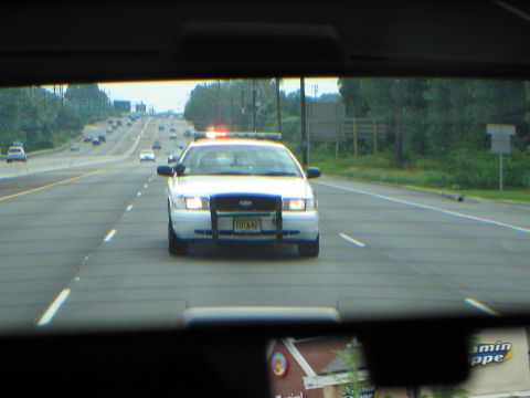 1991 Ford LTD Crown Victoria - white Ford Crown Victoria police car with activated lights follows directly behind, framed by the rear window of the lead vehicle.