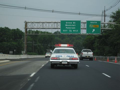 1991 Ford LTD Crown Victoria - white State Police sedan with red light bar travels a highway beneath green signs for Grove St, Montclair and Paterson.