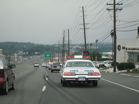 1991 Ford LTD Crown Victoria - white State Police sedan with roof light bar drives along a commercial strip road past a dealership and utility poles.