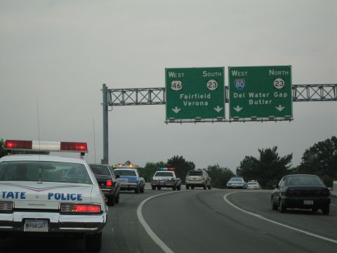 1991 Ford LTD Crown Victoria - Rear view of a white State Police car with red light bar on a curving highway beneath green Route 23, I-80 and Fairfield signs.