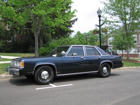 1991 Ford LTD Crown Victoria - Side profile of a dark blue Ford LTD Crown Victoria sedan parked along a curb beside trees and a lamp post.