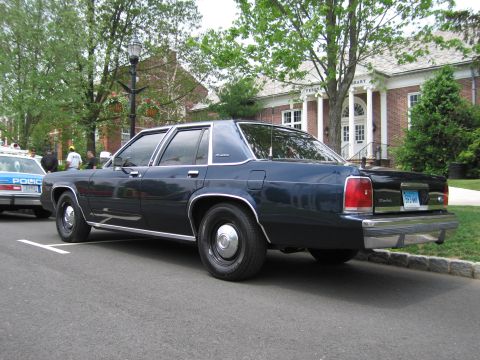 1991 Ford LTD Crown Victoria - Rear three-quarter view of a dark blue Ford LTD Crown Victoria parked at a curb in front of a brick building, a blue police car nearby.