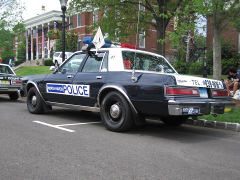 1991 Ford LTD Crown Victoria - Rear three-quarter view of a dark blue North Haven Police Dodge sedan with roof light bar parked at a curb near brick buildings.