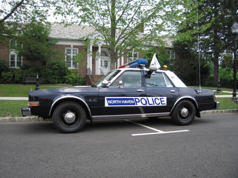 1991 Ford LTD Crown Victoria - Side profile of a dark blue North Haven Police Dodge sedan with light bar and antenna parked along a curb in front of a brick building.