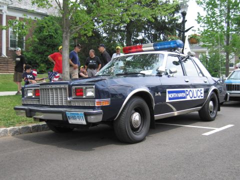 1991 Ford LTD Crown Victoria - Front three-quarter view of a dark blue North Haven Police Dodge sedan with light bar, several people gathered on the sidewalk behind it.