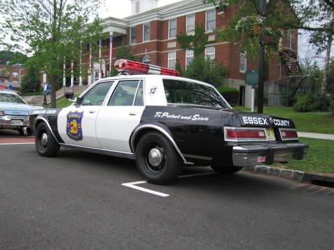 1991 Ford LTD Crown Victoria - black-and-white Essex County police sedan with a roof lightbar parked at a curb in front of a brick municipal building.