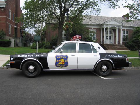 1991 Ford LTD Crown Victoria - Driver-side profile of a black-and-white Essex County Correction Center patrol sedan parked before a brick public library.