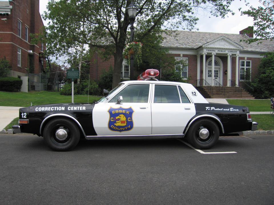 Black and white Essex County Correction Center police car, driver-side profile, in front of brick library