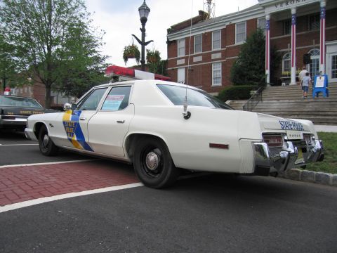 1991 Ford LTD Crown Victoria - white New Jersey State Police Dodge sedan with a blue-and-yellow stripe parked rear three-quarter on red brick pavement.