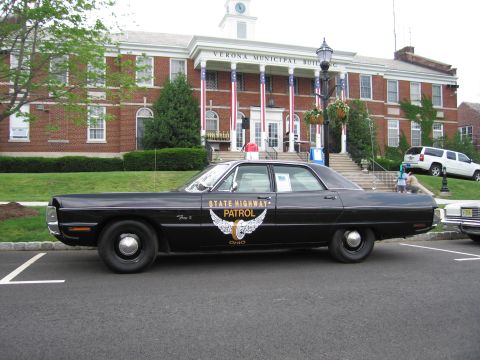 1991 Ford LTD Crown Victoria - Driver-side profile of a black Ohio State Highway Patrol Plymouth Fury with gold winged emblem before a brick municipal building.
