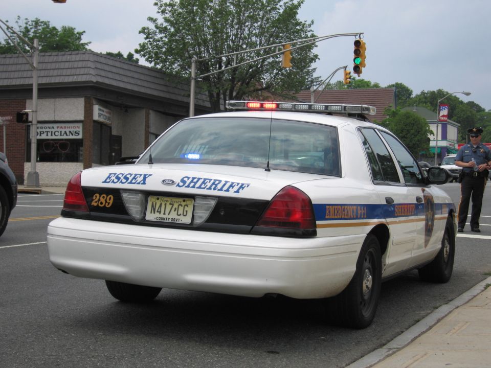 White Essex Sheriff Ford Crown Victoria, rear three-quarter, with lit lightbar at intersection